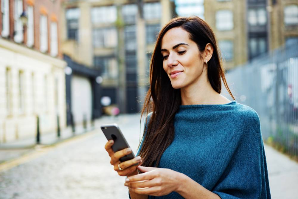 On-page, 2024. A woman stands outside in a city holding her phone and smiling slightly at the screen.