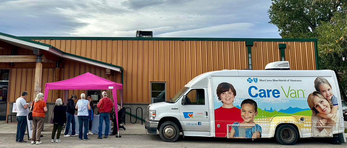 A van with images of children and the Blue Cross and Blue Shield of Montana logo is parked next to a building and a tent where people are lined up for preventive health services. 