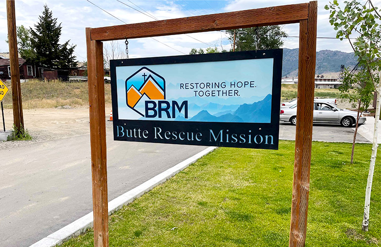 A sign next to a road displays "Butte Rescue Mission" on a blue background with mountains and cross